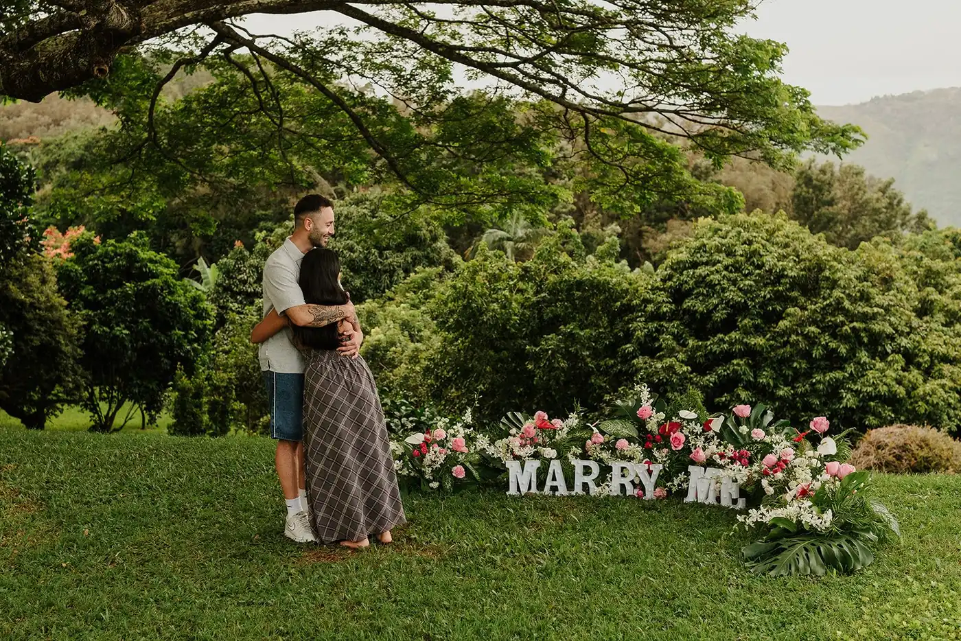 A couple embracing beside a flower-filled Marry Me proposal setup in the Big Island hills